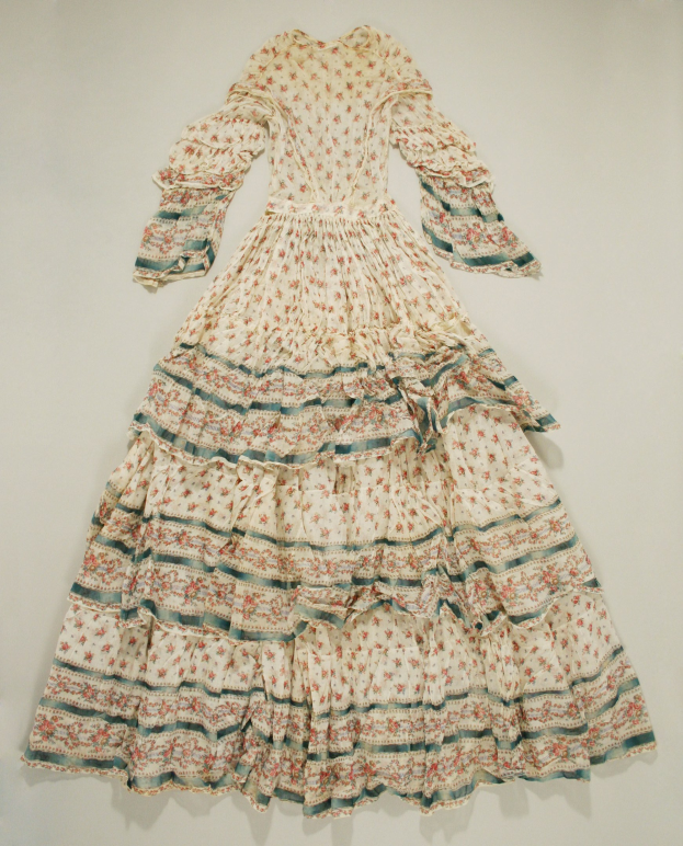 White floral-patterned dress with a fitted bodice, full floor-length skirt, and cascading ruffles hanging on a wall.