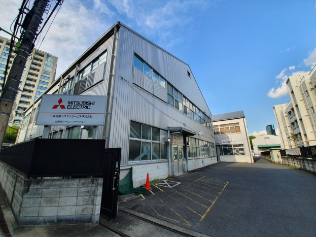 Außenansicht eines Mitsubishi-Electric-Fabrikgebäudes in Tokyo, Japan, mit Glasfenstern, einem Namensschild, Verkehrskegeln, einer Umrandung, einem Tor, einem Strommast mit Drähten, Bäumen und einem bewölkten Himmel.