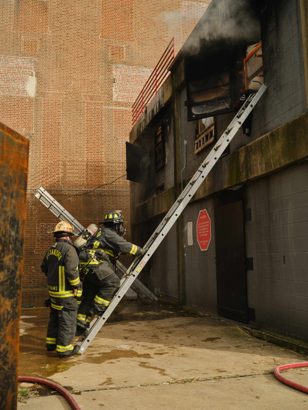 Feuerwehrleute in Helmen, die eine Leiter zu einem rauchgefüllten Gebäude hinaufsteigen, mit einem Metallobjekt links und Rohren am Boden.
