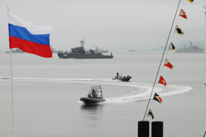 Gruppe von Booten mit Menschen auf dem Wasser mit russischer Flagge im Hintergrund, zusätzliche Flaggen an Stangen rechts und Himmel darüber.