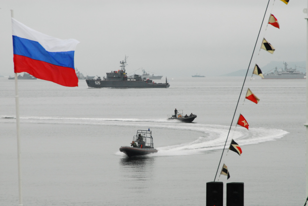 Gruppe von Booten mit Menschen auf dem Wasser mit russischer Flagge im Hintergrund, zusätzliche Flaggen an Stangen rechts und Himmel darüber.