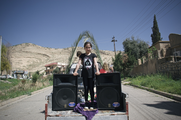 Ein junges Mädchen in einem schwarzen T-Shirt und Hose steht auf einem Lastwagen mit zwei Lautsprechern, die mit einem violetten Tuch bedeckt sind, vor einer Kulisse aus Menschen, Fahrzeugen, Gebäuden, Bäumen, Polen, Drähten, Bergen und einem klaren blauen Himmel.
