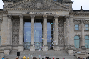 Eine Gruppe von Menschen steht vor dem Reichstaggebäude in Berlin, Deutschland, und spielt mit Seifenblasen, während die Gebäudeplastiken und der Text sowie die Stufen zum Eingang zu sehen sind und der Himmel klar ist.