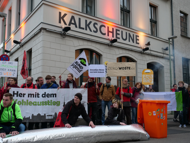 A group of people protesting in front of a building in Germany, holding banners and placards, with two people sitting in the foreground and a dustbin on the right.
