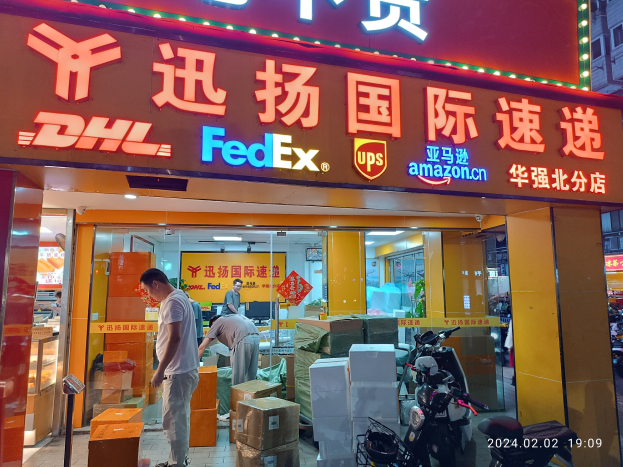 Außenansicht eines DHL- und FedEx-Ladens in Shanghai, China, mit Menschen, Kartons und parkenden Fahrzeugen davor und einem beleuchteten Gebäude im Hintergrund.