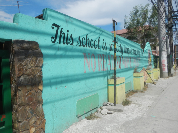 Grüne Wand mit Graffiti, das "Diese Schule ist keine Stadt" liest, umgeben von Gebäuden, Bäumen, Strommasten, Drähten und anderen städtischen Elementen unter einem bewölkten Himmel.