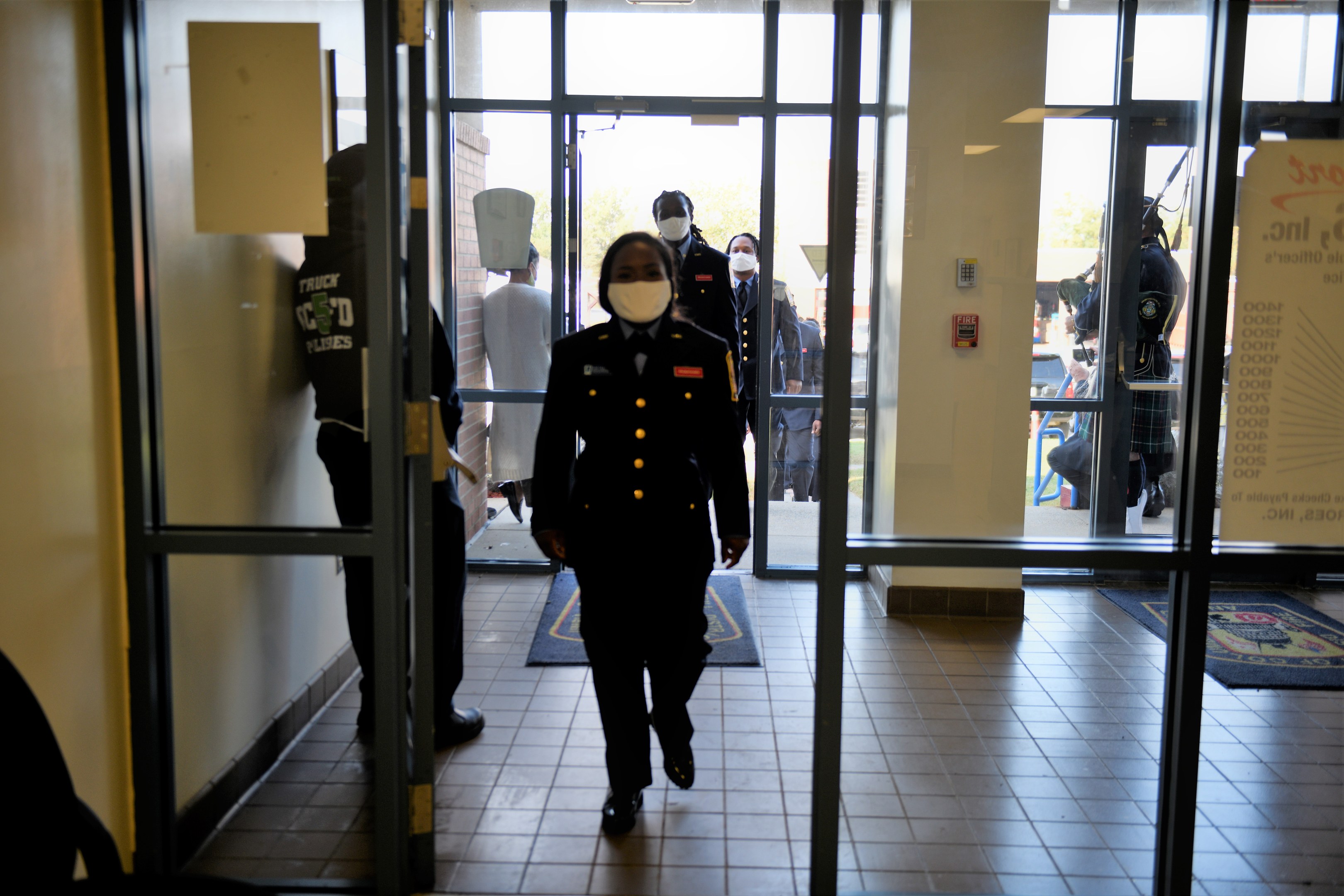 Police officer in uniform and face mask exiting a building with uniformed individuals, a wall-mounted board, glass doors with mats, and outdoor trees visible through the doors.