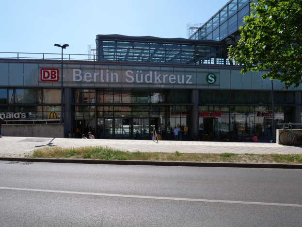 Ein großes Berliner Südkreuz-Bahnhofsgebäude mit Glaswänden und Text, umgeben von Straßenmasten, Straßenlaternen, Kraftfahrzeugen, Menschen auf dem Gehweg, Fahrrädern, Bäumen und einem klaren blauen Himmel.