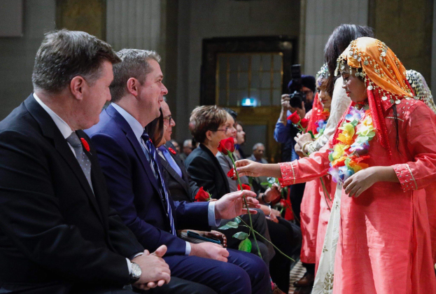 Eine Gruppe von Menschen, die in einem Raum während einer Hochzeitszeremonie in London sitzen, mit einer Frau in einem Kranz und Blumen in der Mitte, einem Mann in einem Anzug rechts von ihr und einer Person mit einer Kamera links von ihr.