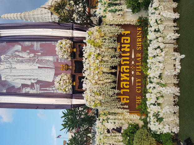 Grand palace in Bangkok, Thailand, adorned with white flowers and greenery, featuring a text board in the foreground and buildings, flags, and trees against a clear blue sky in the background.