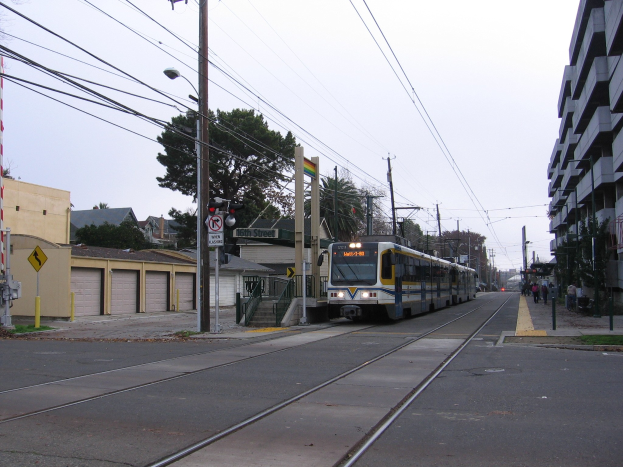 Straßenbahn auf einer Straße zwischen hohen Gebäuden mit Strommasten und Drähten, Fußgängern auf dem Gehweg, Bäumen und einem klaren blauen Himmel im Hintergrund.