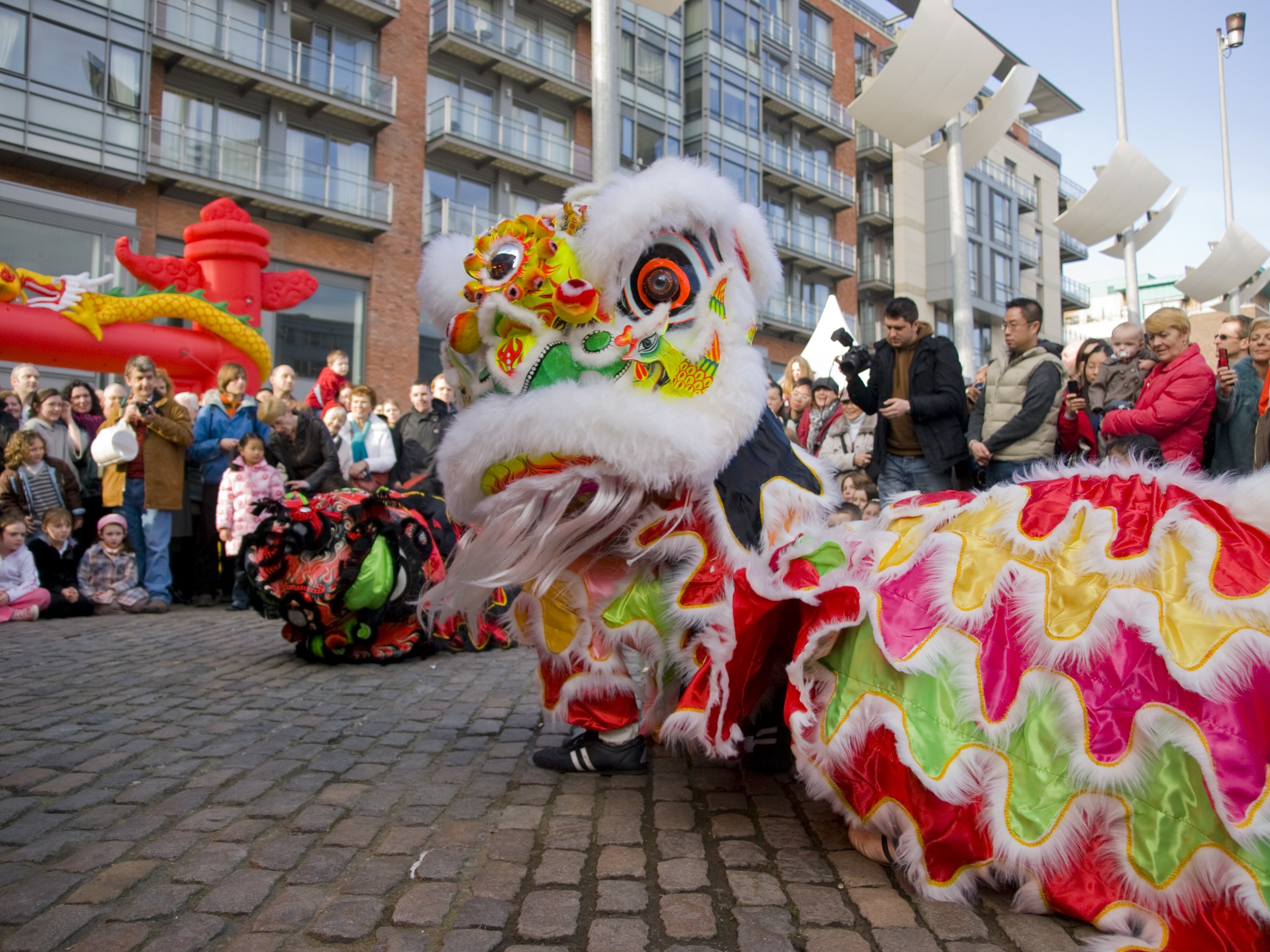 Ein farbenfrohes chinesisches Neujahrsfest in Amsterdam mit einem Löwen tanzen vor einer Zuschauermenge, einige machen Fotos, vor einem Hintergrund aus Gebäuden, Laternenmasten und einem klaren blauen Himmel.