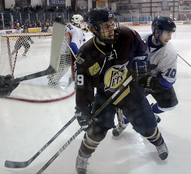 Junge Männer in Helmen und Hockey-Ausrüstung spielen Eisockey auf einer Indoor-Eisfläche mit Zuschauern in den Rängen und beleuchteten Torpfosten im Hintergrund.
