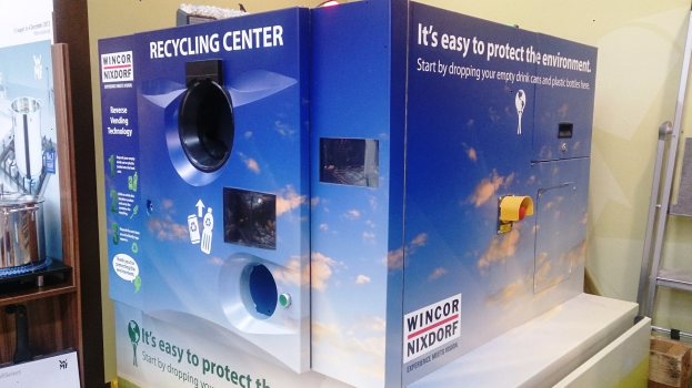 Blue recycling center with a "Recycling Center" sign in the center, a rack of objects on the left, an object on the floor on the right, and lights and a wall in the background.