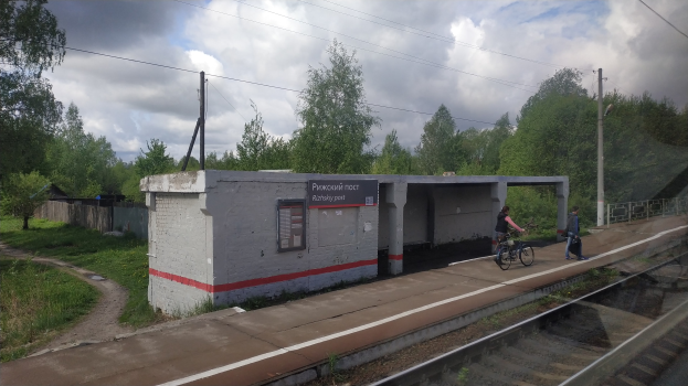 Eine Person, die ein Fahrrad auf einem Bahnsteig neben einer Plattform fährt, mit einem Schuppen, Strommasten mit Drähten, Bäumen und einem bewölkten Himmel im Hintergrund.