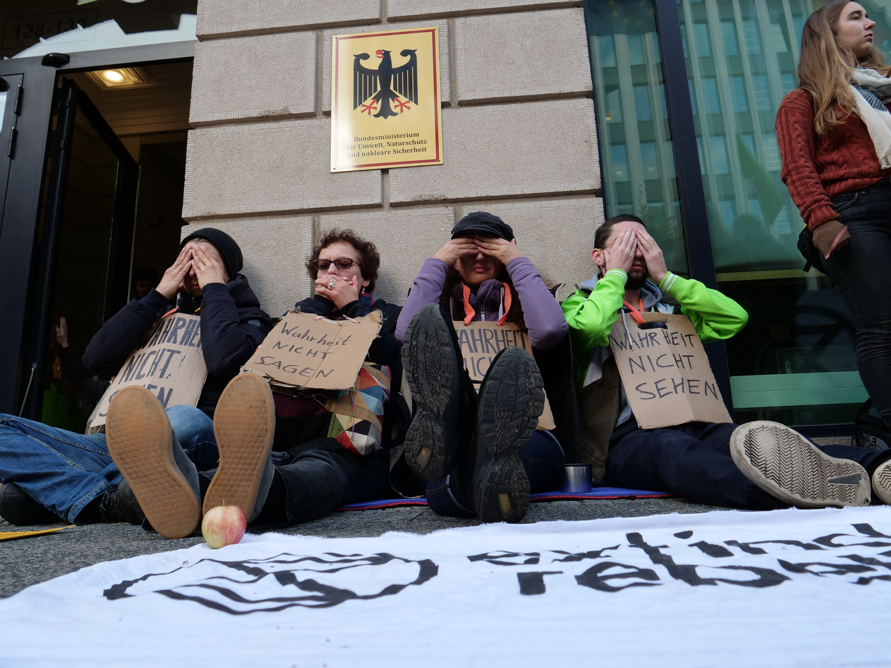 Eine Gruppe von Menschen sitzt vor einem Gebäude auf dem Boden und hält Schilder und eine Fahne mit Text. Eine Frau steht rechts daneben; das Gebäude hat Glasfenster und eine Tür, und in der Ferne ist eine Wand mit einer Tafel zu sehen.