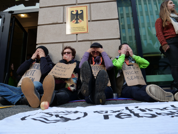 Eine Gruppe von Menschen sitzt vor einem Gebäude auf dem Boden und hält Schilder und eine Fahne mit Text. Eine Frau steht rechts daneben; das Gebäude hat Glasfenster und eine Tür, und in der Ferne ist eine Wand mit einer Tafel zu sehen.