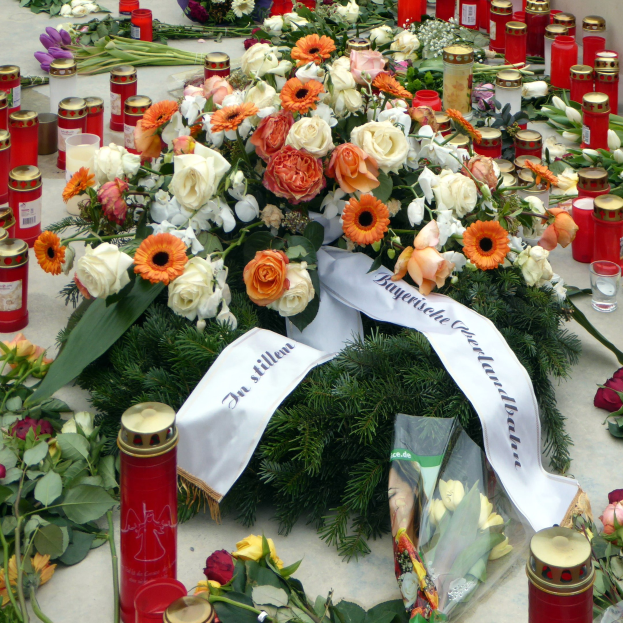 Ein Mahnmal in Berlin, Deutschland, mit einem Blumenstrauß und Kerzen, die auf dem Boden vor einer Wand aufgestellt sind und von Blättern und anderen Gegenständen umgeben sind.