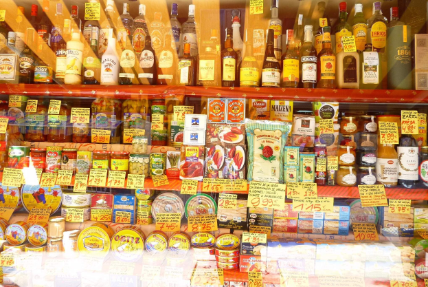 Ein gut sortierter Supermarktgang mit verschiedenen Lebensmitteln und Getränken auf den Regalen, darunter Flaschen, Schachteln und Preisschilder.