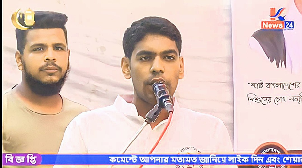 Zwei Männer stehen nebeneinander vor einem Fernseher, einer spricht in ein Mikrofon und der andere trägt ein graues T-Shirt, während sie vor einer Banner mit Text und Logo sprechen.