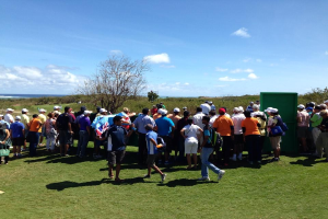 Gruppe von Menschen auf einem grünen Feld bei einem Golfturnier, mit Bäumen, Pflanzen, Wasser und bewölktem Himmel im Hintergrund.