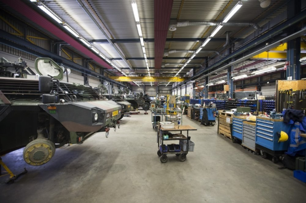 Großes Lagerinnere mit Maschinen, Fahrzeugen links, Transportwagen und Holzboxen rechts sowie Deckenbeleuchtung.