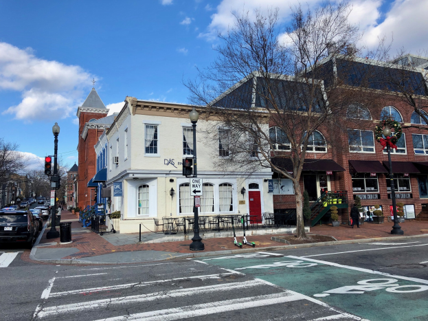 Eine Stadtstraße im historischen Bezirk von Annapolis, Maryland, mit parkenden Autos, Laternenmasten, Verkehrszeichen, Bäumen, Gebäuden mit Fenstern, Außenmöbeln und einem bewölkten Himmel.