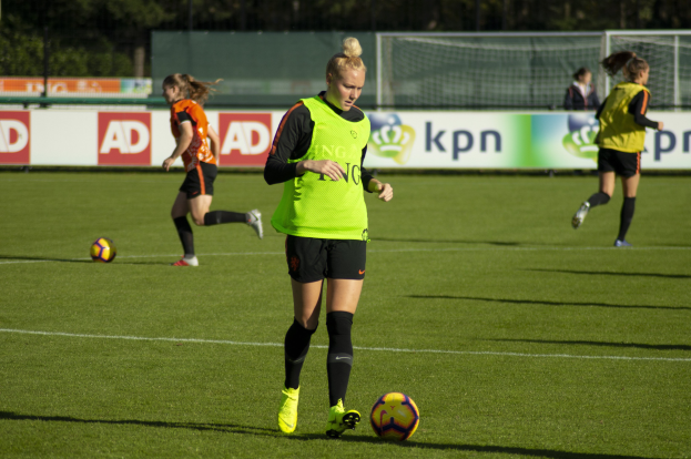 Gruppe von Frauen in Fußballtrikots beim Spiel auf einem Feld mit Bäumen im Hintergrund, Schilder mit Text und ein Netz im Vordergrund.