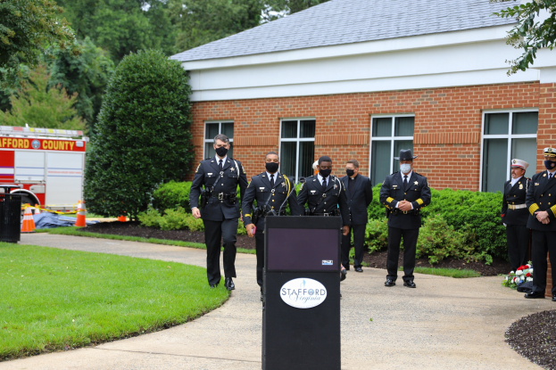Polizisten in Uniform und mit Masken stehen vor einem Gebäude mit einem Podium, Mikrofon und Texttafel im Vordergrund, mit Bäumen, Pflanzen und einem Löschfahrzeug im Hintergrund.