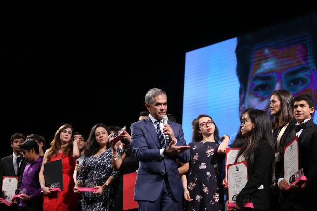 Ein Mann in Anzug und Krawatte hält ein Mikrofon auf einer Bühne, umgeben von Menschen mit Auszeichnungen, mit einem Bildschirm im Hintergrund.