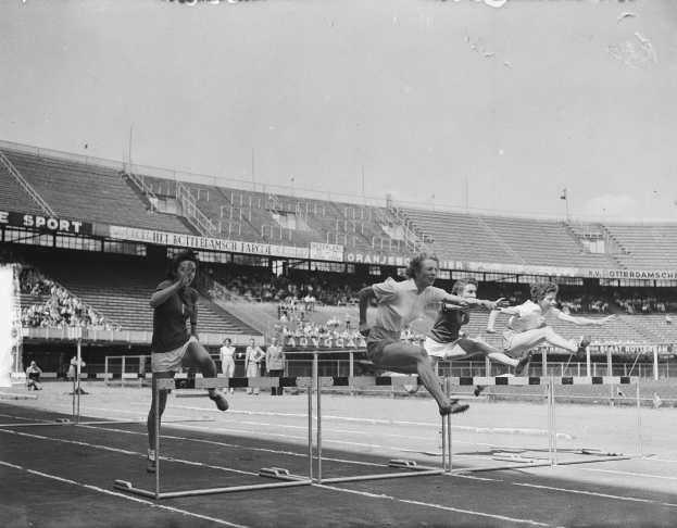 Schwarz-weiß-Foto von Athleten, die Hürden auf einer Laufbahn überspringen, mit Zuschauern auf den Tribünen und einem klaren Himmel.