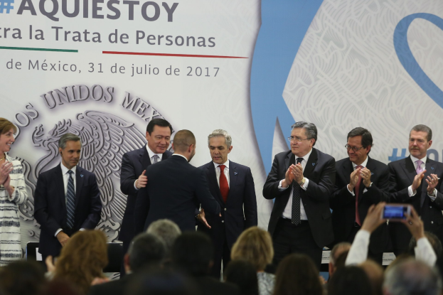 Gruppe von Menschen in Anzügen und legerer Kleidung vor einer Menge, einige klatschen, mit einer Person mit einem Handy im Vordergrund und einem Banner mit Text und einem Bild im Hintergrund.