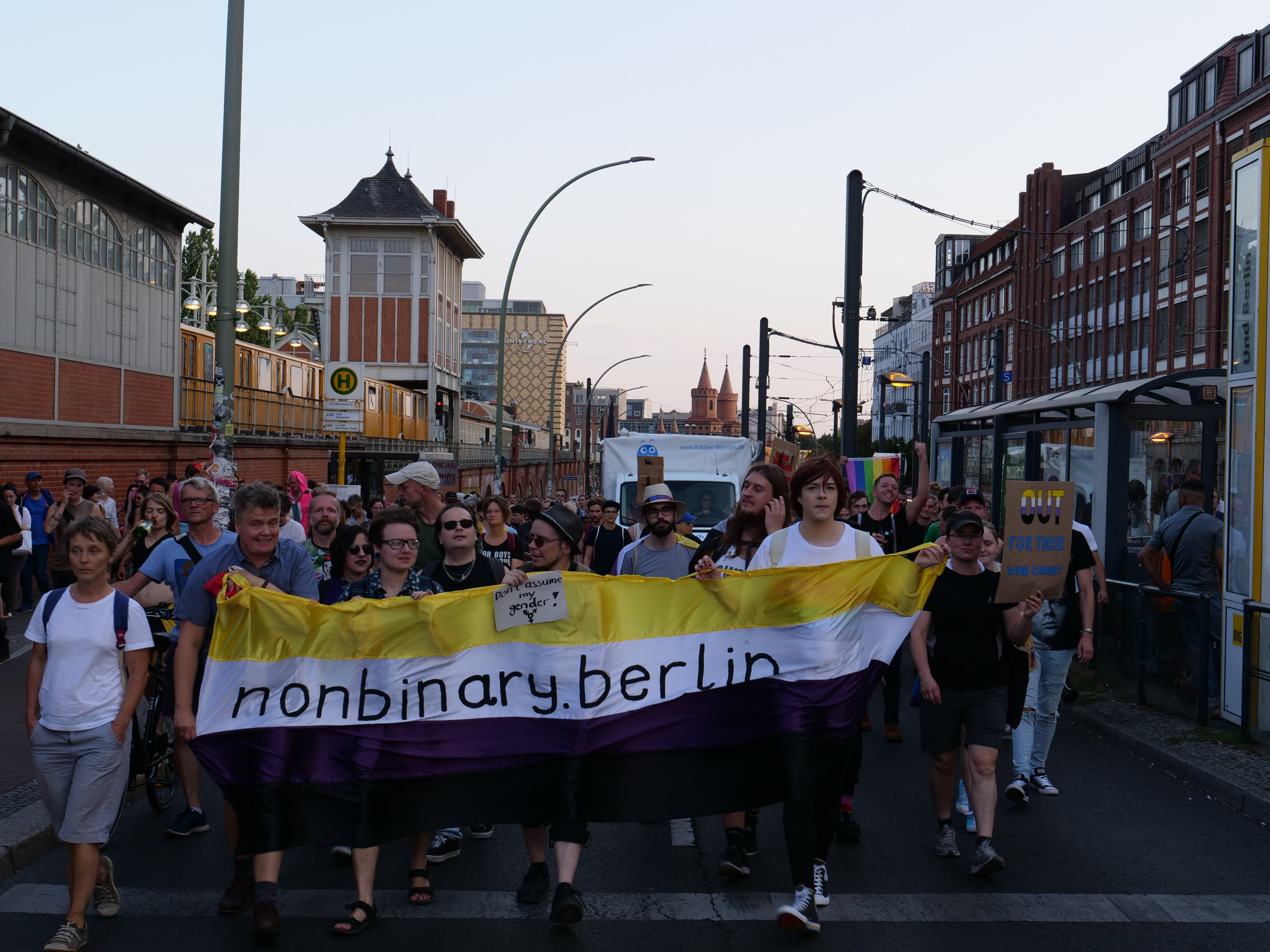Eine Gruppe von Menschen, die eine "Nonbinary Berlin"-Fahne schwingend die Straße entlanggeht, mit Laternenpfählen, Gebäuden, einem Bushaltestellenschild, Bäumen und einem klaren blauen Himmel im Hintergrund.