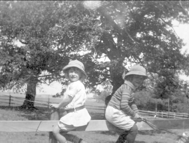 Zwei junge Jungs in Mützen stehen in einem grünen Park und halten Baseballschläger, mit Bäumen und einem Zaun im Hintergrund, auf einem Schwarz-Weiß-Bild.