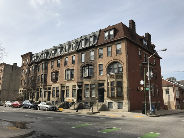 Großes Backsteingebäude an einer Straßenecke mit Fenstern, Treppen, Geländern, Laternen, Schildern, Bäumen und Fahrzeugen, vor einem bewölkten Himmel.