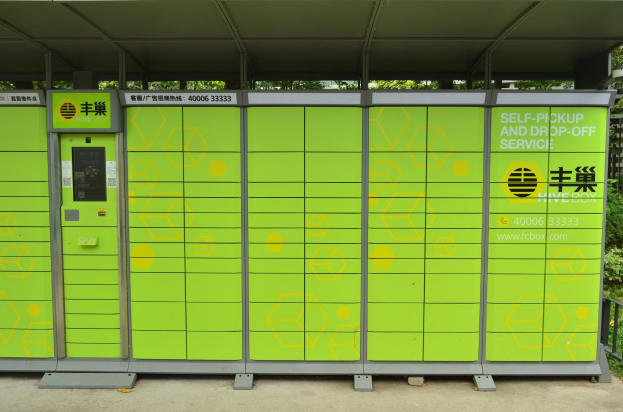 Ein grüner und gelber Verkaufsautomat mit Dach und Text, der die Selbstabholung und -rückgabe anzeigt, steht an der Straße, umgeben von einem Metallzaun, Pflanzen, einer Gruppe von Bäumen und einem bewölkten Himmel.