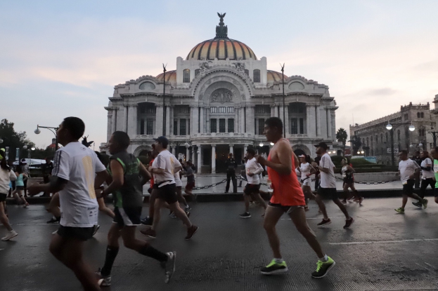 Gruppe von Menschen, die vor einem großen Gebäude während eines Marathons laufen, mit Straßenmasten, Laternen, Bäumen und bewölktem Himmel im Hintergrund.