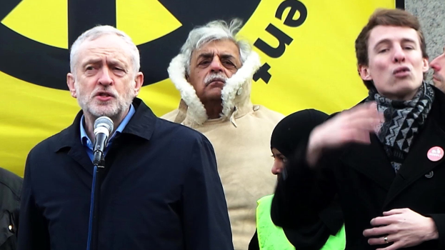 Jeremy Corbyn bei einer Kundgebung in London, stehend an einem Mikrofon mit einer Menge dahinter und einem Banner im Hintergrund.