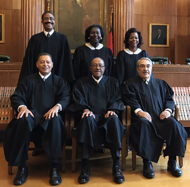 Gruppe von US-Supreme-Court-Richtern in schwarzen Roben, die in einem Gerichtssaal sitzen, lächeln, mit Flaggen, gerahmten Fotos, Deckenleuchten und einem Tisch mit Büchern im Hintergrund.