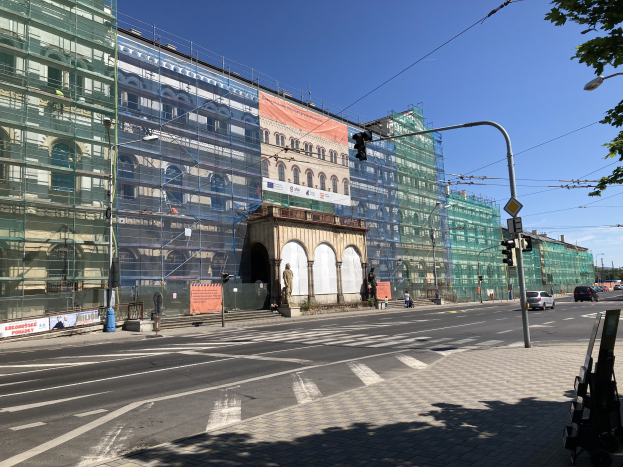 Großer mehrstöckiger Bau in Berlin mit zahlreichen Fenstern, umgeben von Straßeninfrastruktur, Fahrzeugen, Fußgängern und Bäumen unter einem bewölkten Himmel.