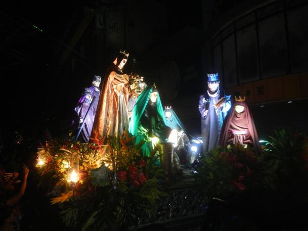 Eine Gruppe von Menschen auf einem geschmückten Paradewagen bei Nacht, umgeben von Pflanzen, mit einem Stadtgebäude im Hintergrund.