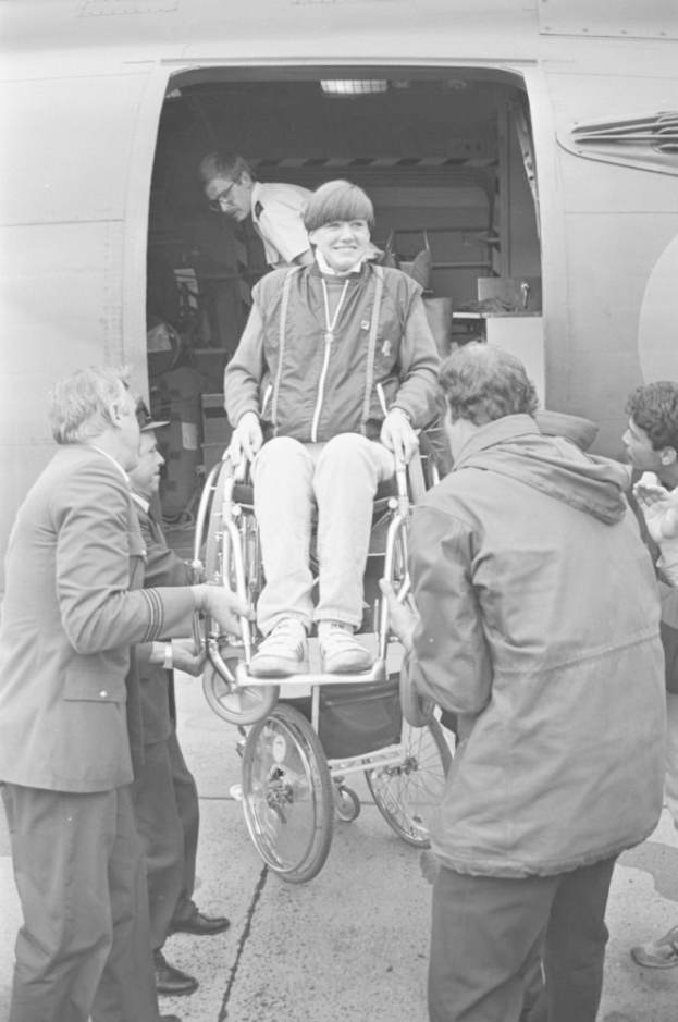 Ein Mann im Rollstuhl wird von einer Gruppe Menschen in ein Flugzeug geholfen, wobei das Flugzeug im Hintergrund zu sehen ist, in Schwarz-Weiß.