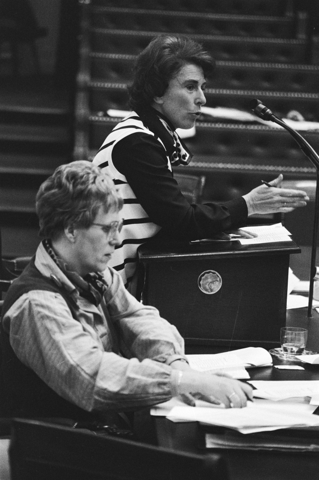 Schwarz-weiß-Foto einer Frau, die an einem Tisch im Europäischen Parlament in Straßburg im Mai 1981 sitzt und einen Stift hält, mit Papieren, einem Glas Wasser und anderen Gegenständen auf dem Tisch, Treppe im Hintergrund.