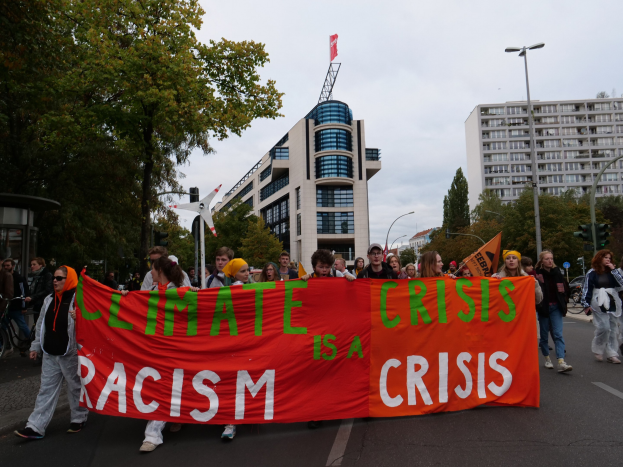 Eine Gruppe von Menschen, die eine Straße entlanggehen und ein Banner mit der Aufschrift "Klima-Krise ist eine Krise" tragen, mit Bäumen, Laternenmasten und Gebäuden an der Straße.