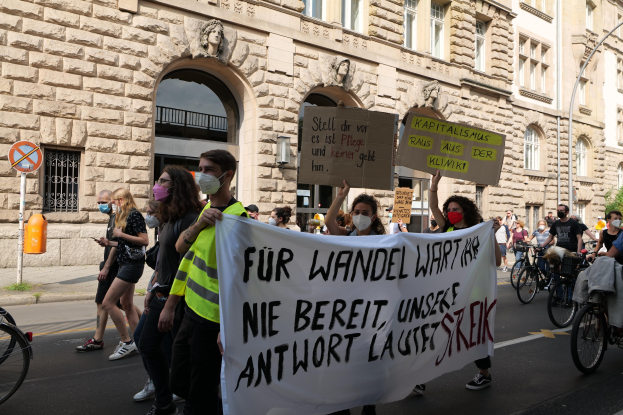 Eine Gruppe von Menschen nimmt an einer Protestaktion gegen die Anti-Kriegs-Bewegung in Berlin teil und marschiert mit Plakaten und Transparenten sowie Fahrrädern die Straße entlang, vor einem Gebäude mit Fenstern, Bögen, Säulen, Skulpturen, Bäumen und einem Laternenpfahl.