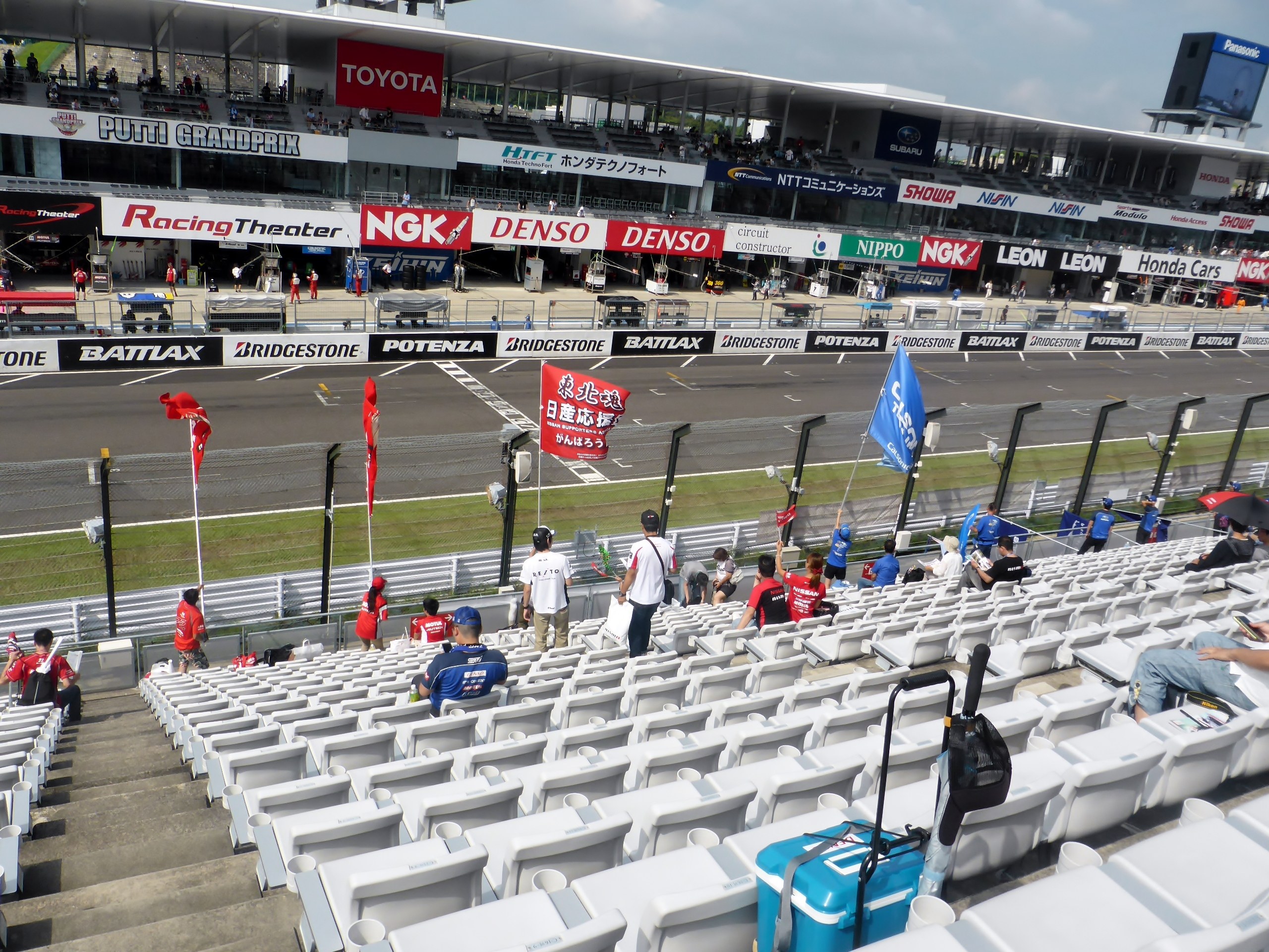 Gruppe von Menschen auf Tribüne einer Rennstrecke mit Fahnen, Schildern, Geländern und Stufen, die auf einen bewölkten Himmel blicken.