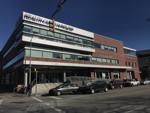 Großes Gebäude mit der Aufschrift 'New York City Health Care Homeless Center' mit parkenden Autos, Fußgängern, Straßenmöbeln und Bäumen unter einem klaren blauen Himmel.