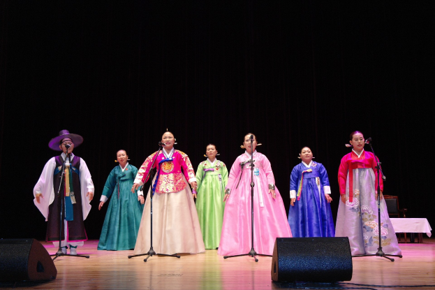 Eine Gruppe von Menschen auf einer Bühne, die in Mikrofone singen, tragen bunte Kleider, mit Lautsprechern vorne und einem drapierten Tisch rechts gegen einen dunklen Hintergrund.