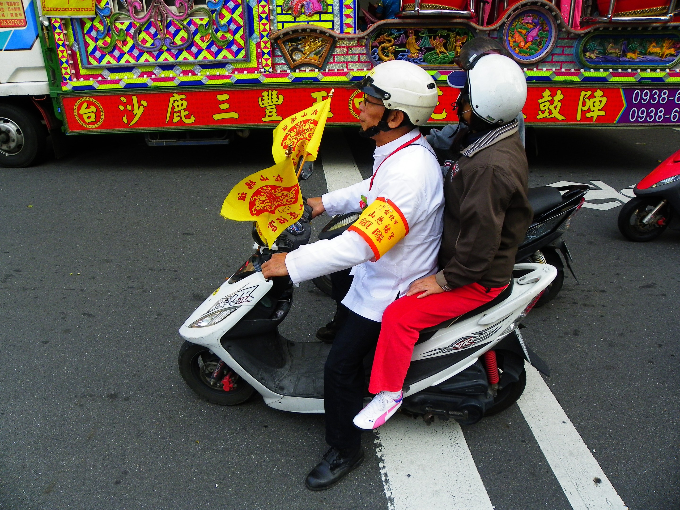 Zwei Personen mit Helmen fahren auf einem Motorroller, eine hält eine gelbe Flagge, mit einem Fahrzeug im Hintergrund.