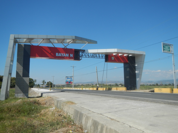 Mäßstelle mit einem 'Bayan N'-Schild an der Seite einer Straße, umgeben von Gras, Strommäasten, Bäumen, Hügeln und einem bewölktem Himmel.